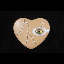 Load image into Gallery viewer, A heart-shaped spell book with a stitched look and green eye design on the front cover, predominantly in a beige color with black and grey accents.
